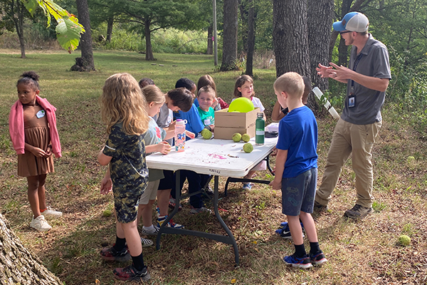 Students stand around a table set up outside looking at items as a teacher stands next to them.